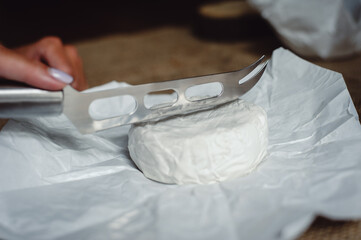 Cut Camembert cheese, French soft cheese with white mold in the hands of a woman on the background of a craft textile tablecloth, close-up. Knife for slicing cheese. Cheesemaker cuts cheese.