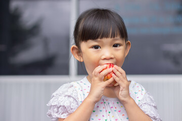 Portrait​ image​ of​ 3-4 yeas​ old​ of​ baby.​ Happy​ Asian child girl eating and biting an red apple. Enjoy eating moment. Healthy food and kid concept.​ Sucking fingers in the mouth.