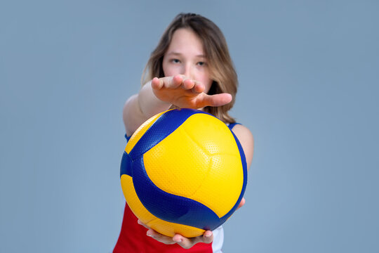 A teenage girl in a sports red volleyball uniform with a ball in her hands stands on a gray-blue background. The hand is ready to hit the ball. The girl is out of focus, Focus on the ball