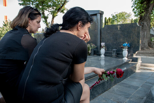 Mother and her daughter in grief, in black clothes, holding a flower and mourning a deceased loved one on cemetery
