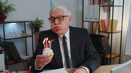 Happy senior mature businessman celebrating lonely birthday in office blowing candles on small cake making a wish. Elderly success grandfather man wearing festive cap celebrate anniversary party alone