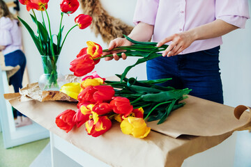 Women's hands pack a festive bouquet in wrapping paper. The florist makes an assembly with red tulips in the workshop. A woman at work, small business or hobby.