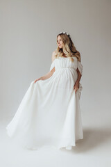 Blonde pregnant woman in a white dress with a wreath on her head in the studio on a white background holds her hand on her stomach