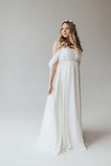 Blonde pregnant woman in a white dress with a wreath on her head in the studio on a white background holds her hand on her stomach