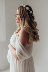 Blonde pregnant woman in a white dress with a wreath on her head in the studio on a white background holds her hand on her stomach