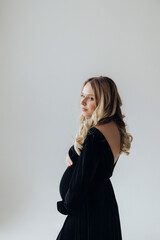 Fototapeta premium a pregnant blonde woman in a black dress stands in the studio on a white background holds her hand on her stomach