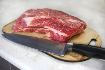 Large fresh piece of meat on a wooden cutting board with a metal knife on a light kitchen table