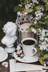 A beautiful postcard. A white coffee cup with a saucer, a statuette, candles, a book and a vase with a bouquet of blooming apple trees. Beautiful still life. Spring time. The concept of 