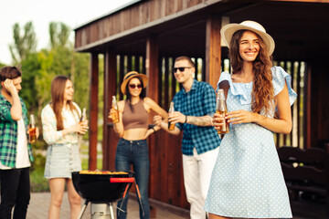 happy beautiful young woman in a hat with lemonade in the foreground, with a group of friends having a barbecue outdoors