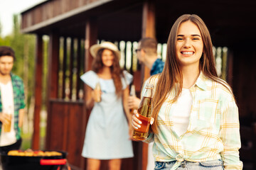 happy beautiful young woman with lemonade in the foreground, with a group of friends having a barbecue outdoors