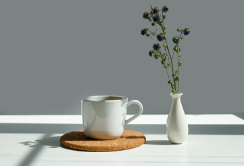 tea on a stand in a white mug with a vase of flowers on a white background in sunlight.