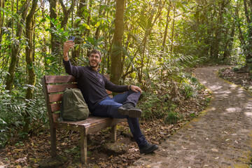 Fototapeta premium Funny Caucasian man taking a selfie in nature. He is sitting next to a stone path. He is wearing hiking clothes and backpack. Costa Rica, Monteverde.