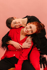Two pretty caucasian young ladies are leaning on each other, sitting on pink background. Brunette and redhead are wearing red, black suits. Сoncept lifestyle sincere emotions of people.