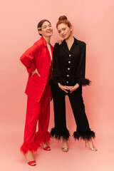 Full-length beautiful young caucasian women brunette and redhead look at camera on pink background. Models wear elegant suits and heeled shoes. Stylish ladies having good time.
