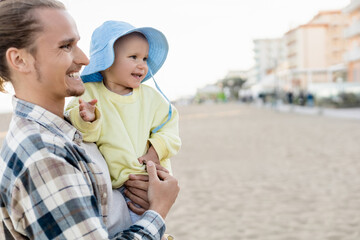 Fototapeta premium Smiling father and baby girl looking away on beach in Italy.