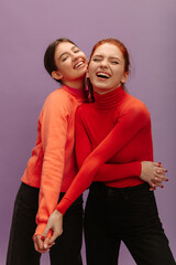 Cheerful young caucasian women in good spirits hugging on purple background. Brunette and redhead are wearing sweater with pants. Lifestyle, different emotions, leisure concept. 