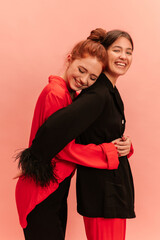 Happy young caucasian girls embrace each other standing one by against pink background. Red-haired model with eyes closed leans on shoulder of brunette wearing pajama-style suit.