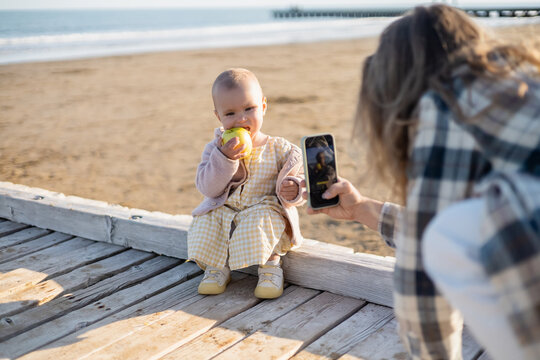 Blurred father taking photo of baby with apple on wooden pier in Italy.