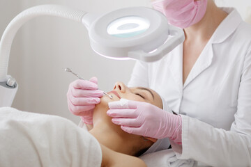 Young woman gets facial cleansing in a cosmetologist's office. Skin cleaning at beauty salon. Beautician squeezing pimples and blackheads from face.