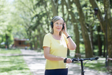 Senior happy woman on a walk in the park with a bicycle listens to music on headphones, and walks on a summer day