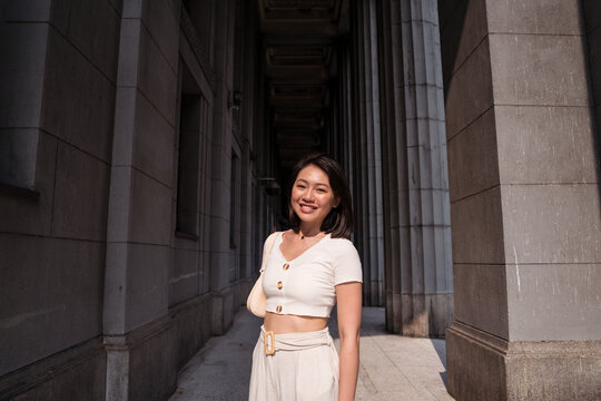 Content young Asian lady standing near building with columns in sunlight