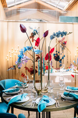 in the dark hall there are festive tables and chairs, tables decorated with compositions of flowers, greenery and candles. On the tables are glasses, plates and cutlery