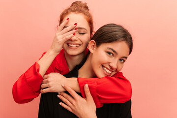 Close-up of young caucasian girl with her eyes closed hugging smiling friend from back against pink background. Brunette and redhead are wearing casual clothes. Concept of lifestyle emotions people.