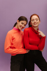 Obraz premium Modern young caucasian girls wearing sweater and pants pose on purple background. Brunette looking at camera, redhead with her eyes closed. Mood, lifestyle, concept .