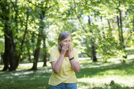 An elderly woman in the park with allergies has a runny nose and sneezes near trees