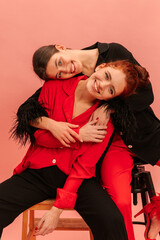 Two cute young caucasian girls are smiling looking into camera with heads on each other against pink background. Brunette and redhead wear elegant suits for photo shoot. Lifestyle, beauty concept