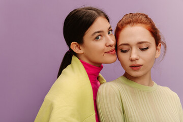 Close-up of two young caucasian women in sweaters snuggling up to each other on purple background. Brunette looks at camera, redhead down. Lifestyle, leisure concept. 
