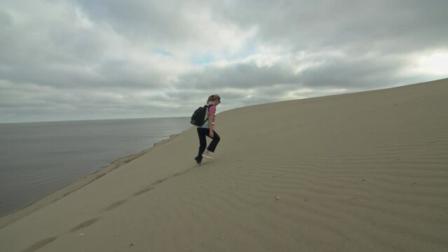 Blonde girl walking on top of dune in jacket. Stylish woman having walk barefoot in desert. Young lady gesturing hand in slow motion, 4k footage