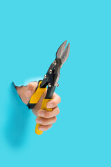 Man hand holding straight cut pliers suitable for cutting plastic or metal materials that breaks through blue wall color background. Aviation snips.