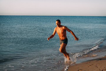 Fototapeta premium A slender muscular tanned guy in blue swimming trunks runs along the sandy shore of the ocean and smiles on a bright sunny day.