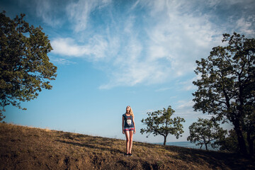 An attractive blonde girl in striped shorts and a blue t-shirt stands on the hills, around the trees, on a bright sunny day. There are many beautiful clouds in the sky.