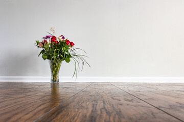A bouquet with roses and decorative long grass leaves placed on the hardwood floor of empty domestic room with white wall