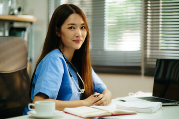 Medium close up portrait of Asian female doctor or nurse with stethoscope smiling toward to camera while sitting in the room. Optimistic medical people on duty. Portrait of medic happiness.