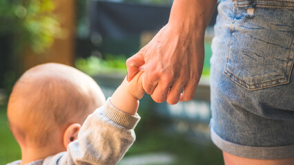 Learning to walk concept: Toddler is holding the hand of his mother, making his first steps. Close up from behind. - 507130536