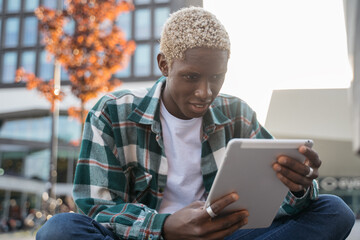 Obraz premium Young smiling African American student using digital tablet studying, learning language sitting in university campus, education concept. Hipster guy with stylish hair communication online outdoors 