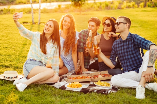 Happy friends have a picnic in the city park outdoors. Friends have fun talking and taking selfie on mobile phone