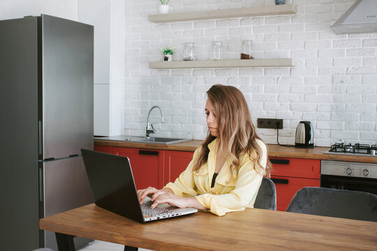 A Young Beautiful Caucasian Blonde Woman In A Yellow Shirt Sitting At The Table Working On A Laptop From Home In The Kitchen. Freelance