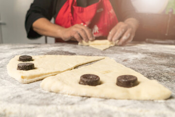 Close-up of pieces of rolled croissant dough with pieces of chocolate on a table sprinkled with flour, in the background a woman cook twists croissants. Selective focus