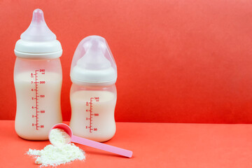 Bottles of different sizes with milk formula and dry baby milk powder in a measuring spoon on a red background. Artificial feeding of the infant. Space for the text.