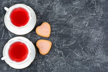Obraz na płótnie Canvas Two cups of raspberry tea on saucers and two beautiful heart-shaped cookies on a black background. Space for the text. Valentine's day, birthday, anniversary. Romantic breakfast.