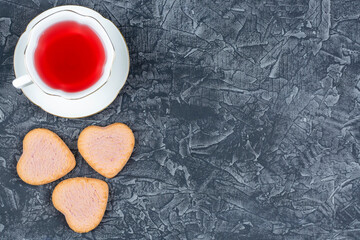 Elegant cup of raspberry tea on a saucer and rich strawberry cookies in the shape of a heart on a black (dark) background. Space for the text. Good morning. The concept of love, loyalty.