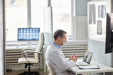 Mature Caucasian radiologist wearing white lab coat sitting at desk in front of laptop working with...