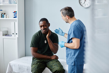 Obraz premium Young Black soldier sitting on medical couch in modern military hospital telling doctor about pain in neck