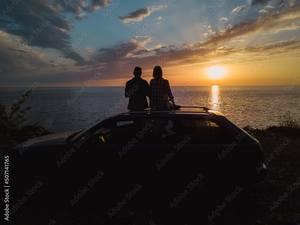 Wall mural Silhouette of a happy couple sitting on the roof of a car on a cliff in front of the sea with a charming beautiful orange-blue sky at sunset, view from a drone. Concept of vacations and travel,romance