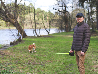 Young millennial male is walking with his cute Japanese breed shiba inu dog puppy on a riverside. Dog assistant emotional support training.