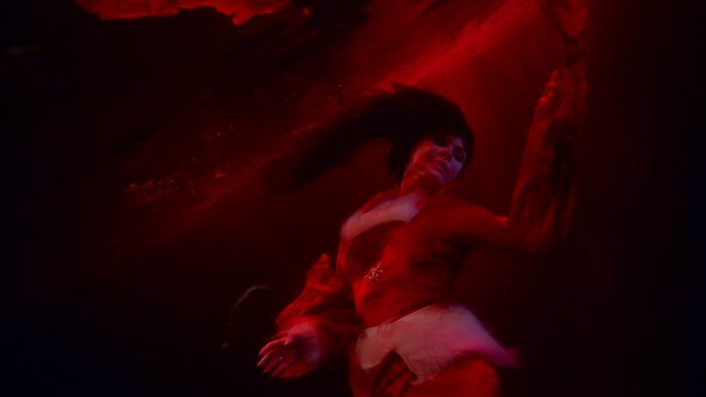 underwater witch in red suit is floating in dark red depth of sea or river, swaying her black hair